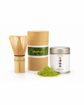 Produktaufnahme mit Bambusbesen, Matcha Society Dose, Seisho Ceremonial Matcha Dose und einem Häufchen feinem Matcha-Pulver vor hellem Hintergrund.