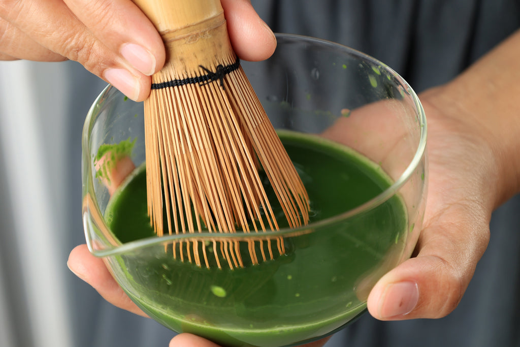 Nahaufnahme eines Bambusbesens, der frisch angerührten Matcha in einer Glasschale schaumig aufschlägt.