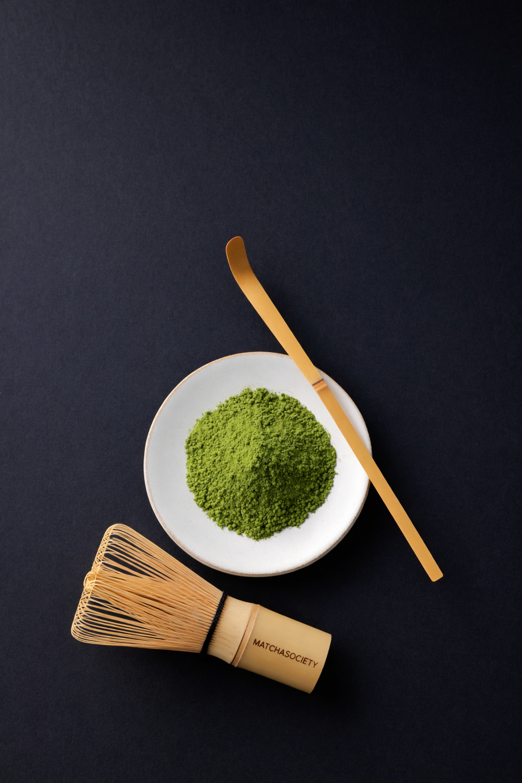 Minimalistische Flatlay-Aufnahme mit Matcha-Pulver in einer weißen Schale, Bambuslöffel und Matcha-Besen auf dunklem Hintergrund.